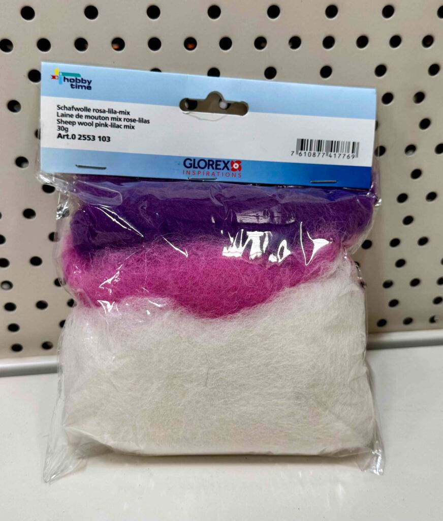 Die getestete Filzwolle in Weiß, Rosa und Lila steht innerhalb der 30-g-Packung auf einem weißen Regal.