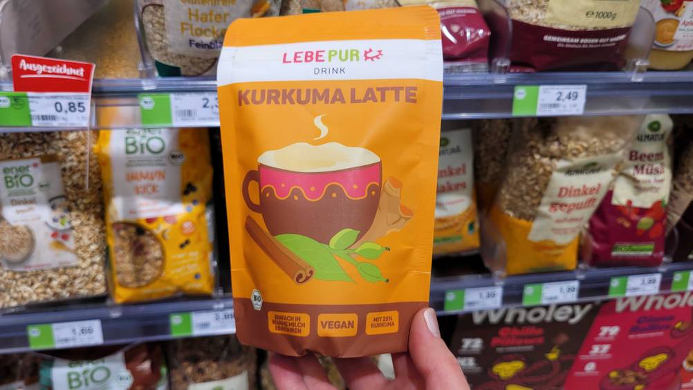 Eine Hand hält eine Packung des getesteten Goldene-Milch-Pulvers vor ein Regal.
