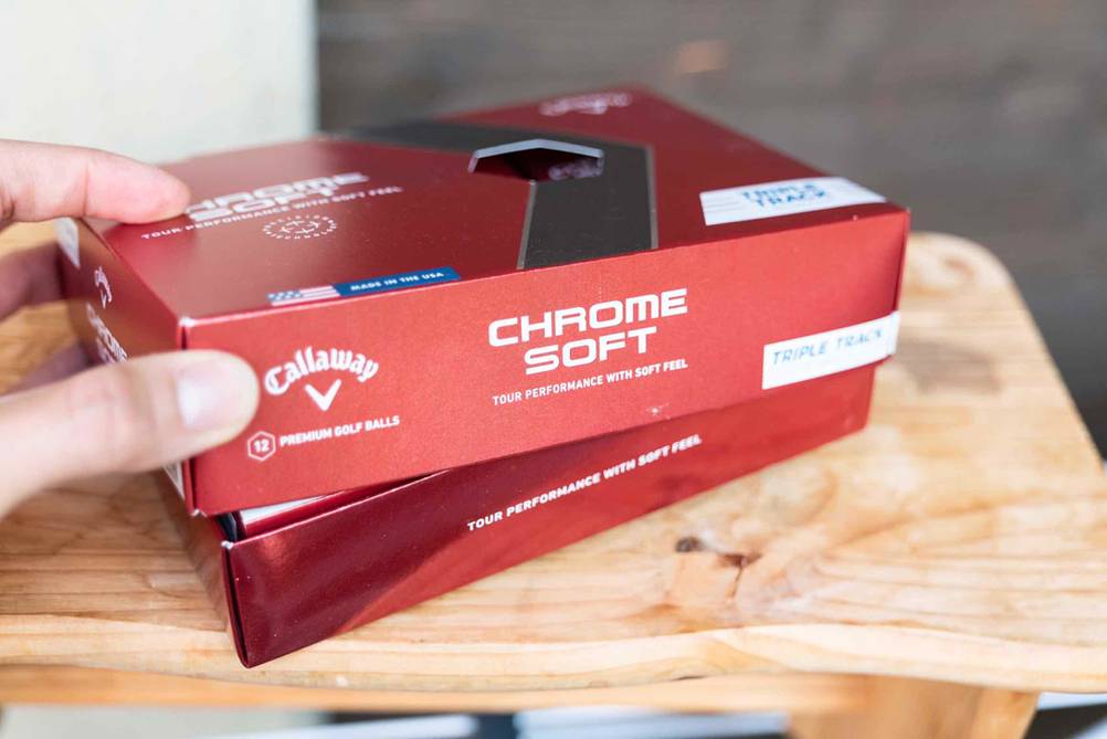 Eine Hand öffnet eine Packung mit getesteten Golfbällen die auf einer Holzbank steht.