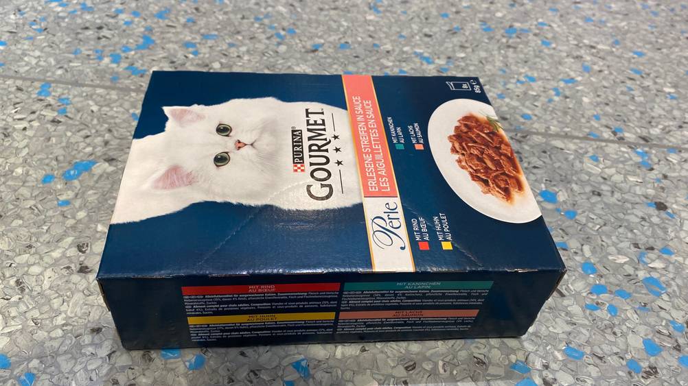 Eine Packung des getesteten Gourmet-Katzenfutters auf der Seite liegend auf einer grauen Fläche.