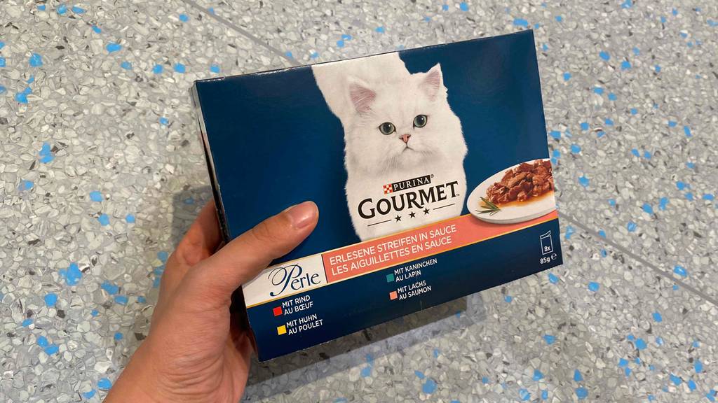 Getestet: Hand hält einen Karton mit 8 Beuteln Gourmet Katzenfutter in verschiedenen Geschmacksrichtungen über einem gesprenkelten Fliesenboden.