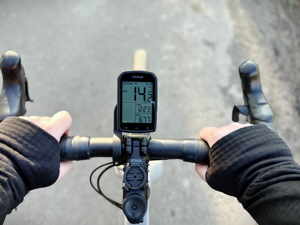 Der Cycplus M1 Fahrradcomputer zeigt während der Fahrt Geschwindigkeit und Distanz an. Die Hände des Fahrers greifen den Rennradlenker.