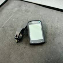 Der Cycplus M1 Fahrradcomputer liegt neben seiner Halterung auf einer dunklen Oberfläche. Das Gerät hat ein großes Display mit drei Bedientasten.