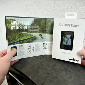 Verpackung des Wahoo ELEMNT BOLT V2 mit geöffnetem Karton und einer Produktbroschüre. Zwei Hände halten die Verpackung und die Broschüre in einem Innenraum.