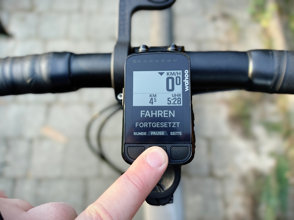 Ein Finger drückt den Pause-Knopf des Wahoo ELEMNT BOLT V2 Fahrradcomputers, der am Lenker eines Rennrads montiert ist. Das Display zeigt die aktuelle Geschwindigkeit und Distanz an.