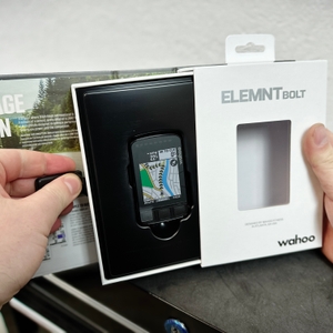 Der Wahoo ELEMNT BOLT V2 in geöffneter Verpackung, gehalten von zwei Händen. Im Hintergrund ist eine Produktbroschüre mit Informationen zu sehen.