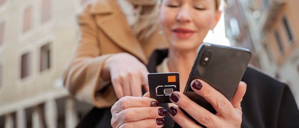 Eine Frau steht hinter einer anderen Frau und deutet auf die in der Hand der vorderen Frau befindlichen SIM-Karte.