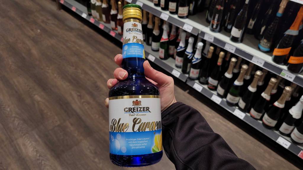 Eine Hand hält eine Flasche mit Blue-Curacao im Test vor einem Getränkeregal im Supermarkt.