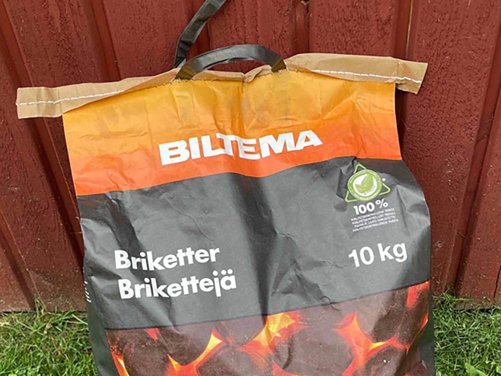 Eine Packung der getesteten Grillbriketts lehnt an einem roten Zaun.