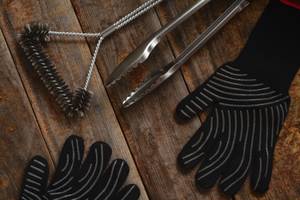 Grillbürste liegt mit Handschuhen und Zange auf einem Tisch.