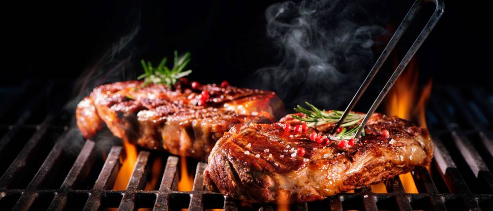 Zwei Steaks mit Rosmarin und Beeren liegen auf dem Grill und werden mit einer Grillgabel aufgespießt.