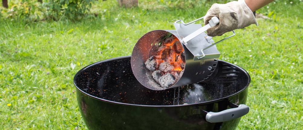 Eine Person mit einem Grillhandschuh kippt glühende Kohle aus dem Anzündkamin in einen Kugelgrill.