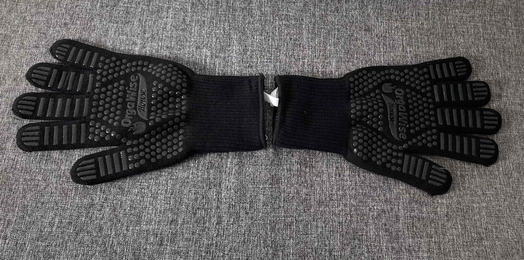 Schwarze getestete Grillhandschuhe liegen mit der gummierten Innenseite nach oben auf einer grau melierten Oberfläche.