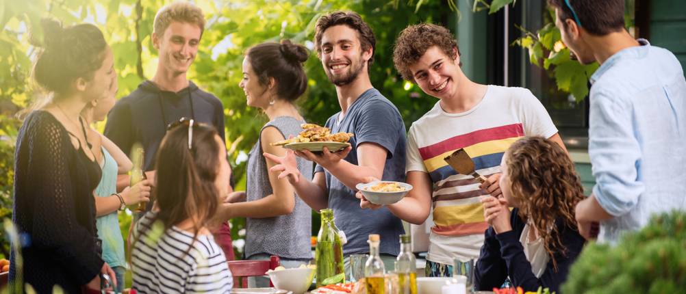 Eine Gruppe junger Freunde steht und sitzt bei einer Grillparty um einen Tisch.