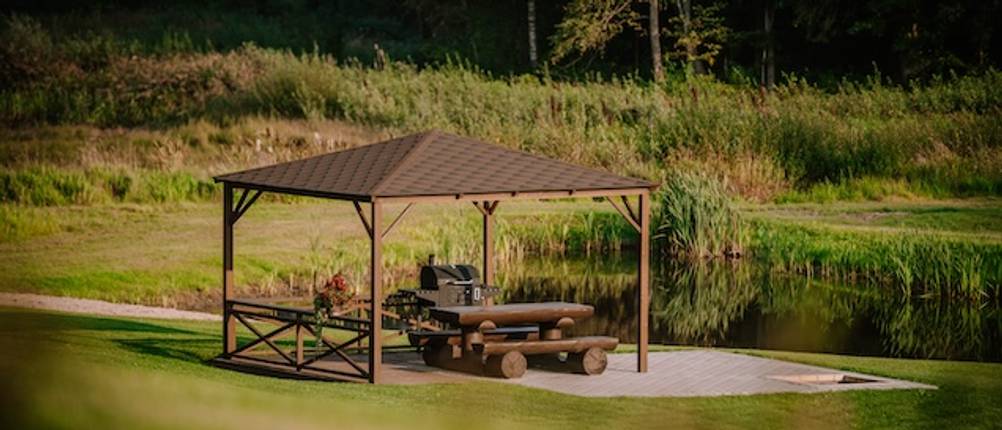 Grillpavillon im Test: Auf einer großen Wiese steht ein Grillpavillon aus Holz mit einer Sitzgarnitur aus Baumstämmen und einem Grill.