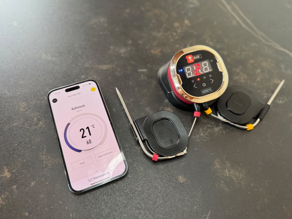 Das getestete Grillthermometer von Weber liegt mit einem Handy und geöffneter Weber-App auf einem Tisch.