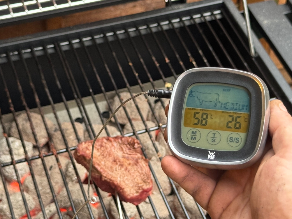 Eine Person hält getestete Grillthermometer von WMF vor einem Grill mit einem Steak in der Hand.
