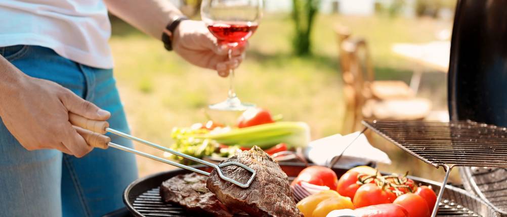 Mann mit einem Glas Wein wendet mit einer Grillzange ein Steak auf einem Grill mit Steaks und Grillgemüse.