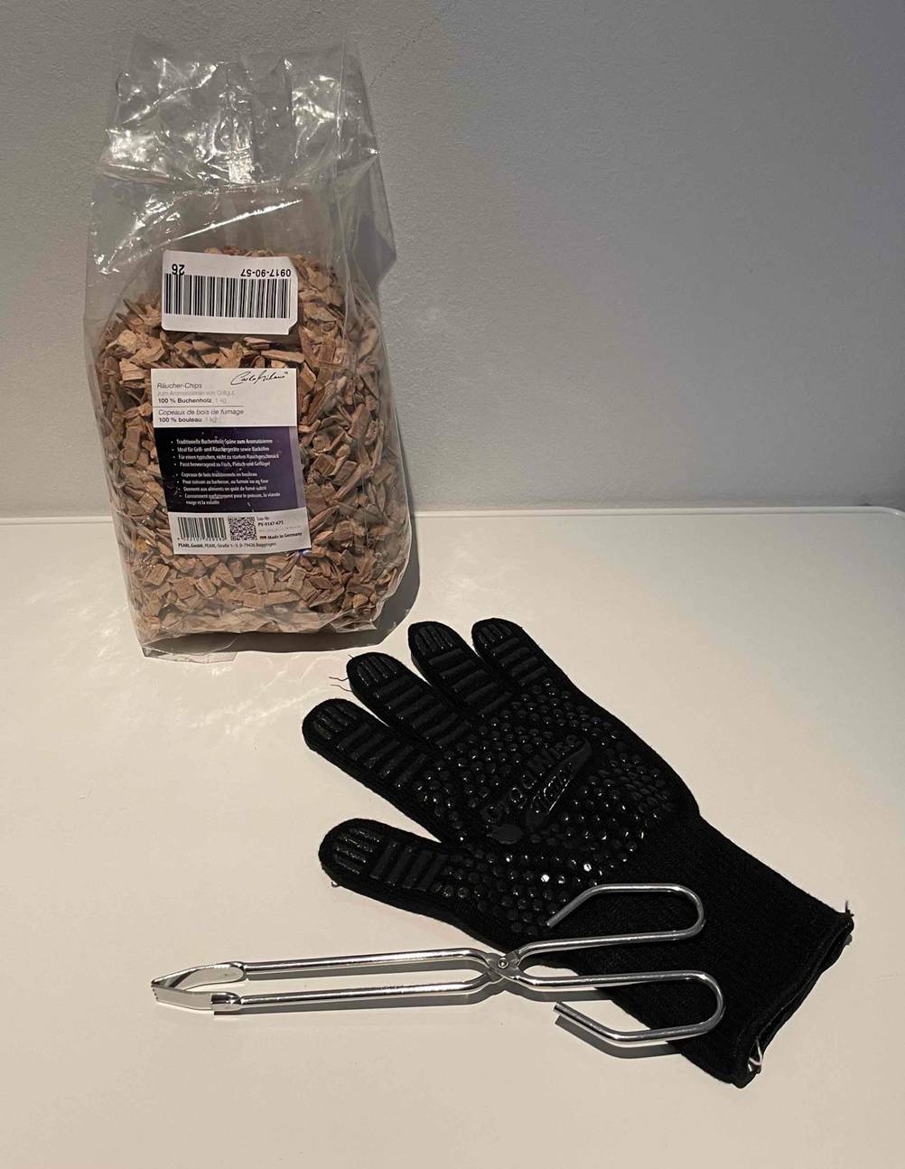 Eine getestete Grillzange liegt quer auf einem Grillhandschuh vor einer Packung Räucherchips.
