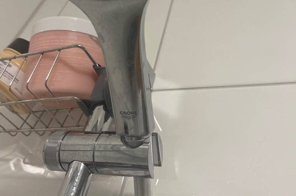 Im Test: Duschkopf von GROHE inklusive Anti-Kalk-Funktion an einer Duschkopfhalterung auf weißer Fliesenwand neben Badeutensilien in einem Metallkorb.