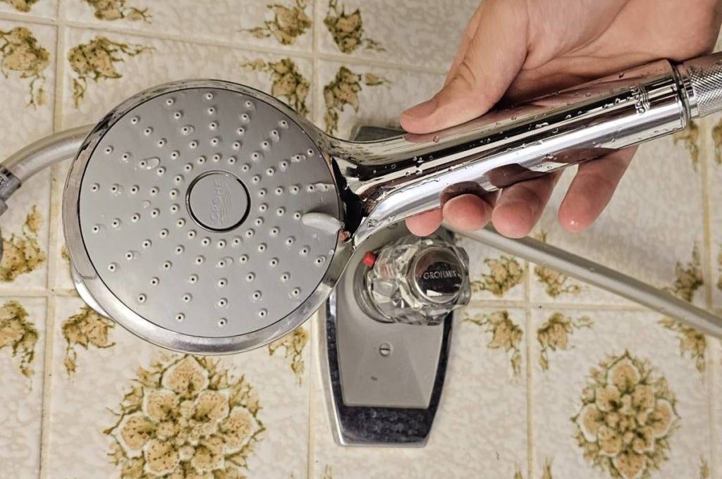 Hand hält einen GROHE Duschkopf im Test vor eine Fliesenwand im 70er-Jahre Design, auf der sich ein GROHE Warmwasser-Regler befindet.