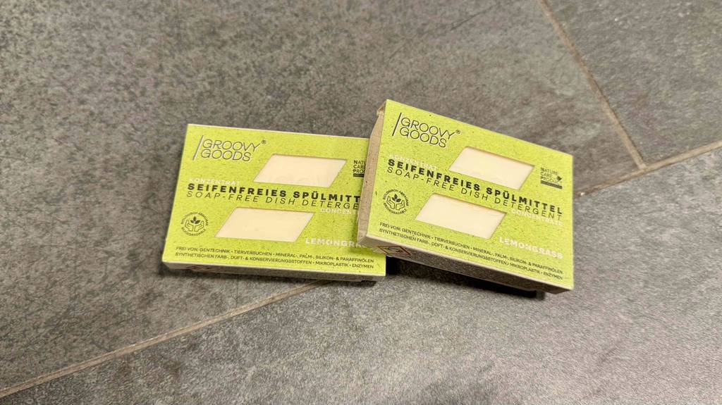 Ein seifenfreies, festes Spülmittel im Test liegt in doppelter Ausführung innerhalb seinen grünen Verpackungen auf einem grauen Fliesenbelag.