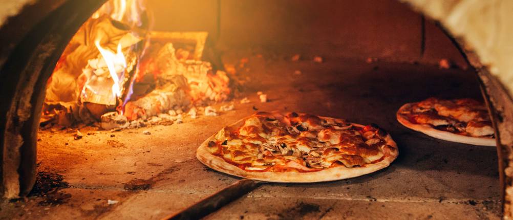 Eine große Pizza wird mit einer Pizzaschaufel in einen Pizzaofen geschoben.