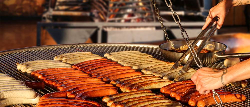 Viele Würstchen werden auf einem Schwenkgrill zubereitet und gewendet.