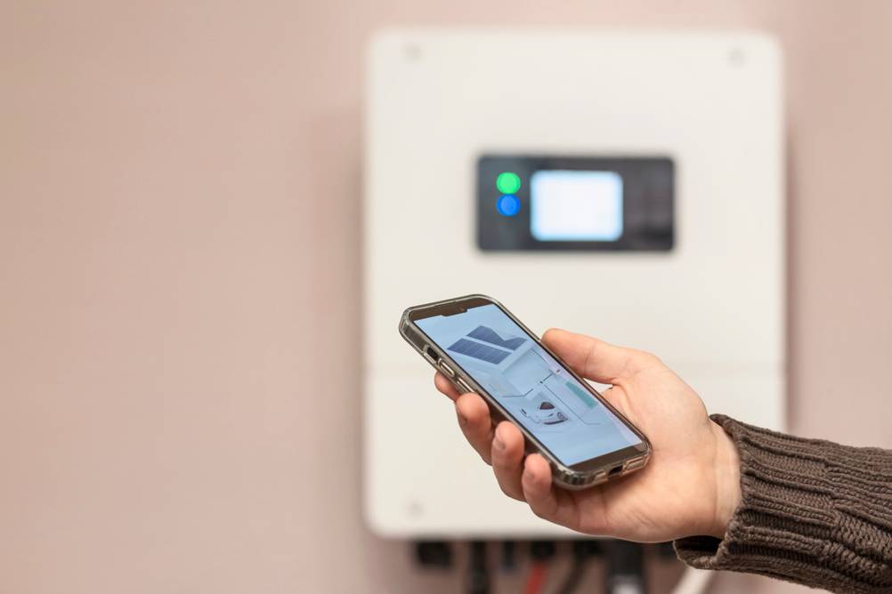 Growatt-Wechselrichter im Test: Eine Hand hält ein Handy auf dem eine schematische Anlage mit E-Auto, Wallbox und PV-Anlage zu sehen ist vor einem diffus sichtbaren Wechselrichter.