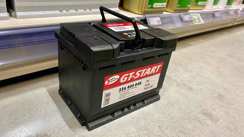 Eine Autobatterie im Test von leicht seitlich mit hochgeklapptem Griff auf einem Betonboden vor einem Verkaufsregal.