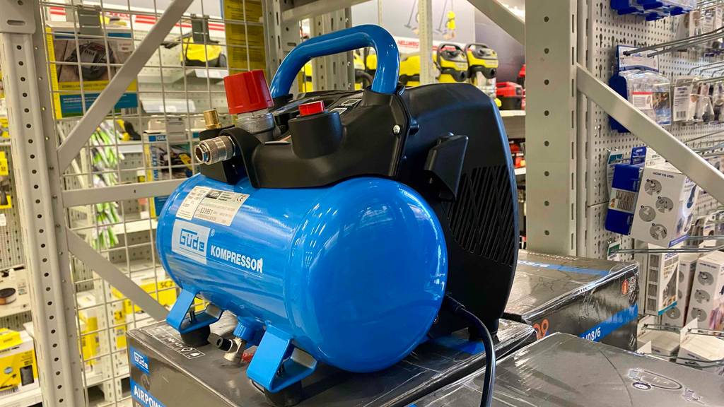 Seiten- und Vorderansicht auf einen blau-schwarzen Güde Kompressor im Test, der auf seiner Originalverpackung in einem Verkaufsbereich steht.