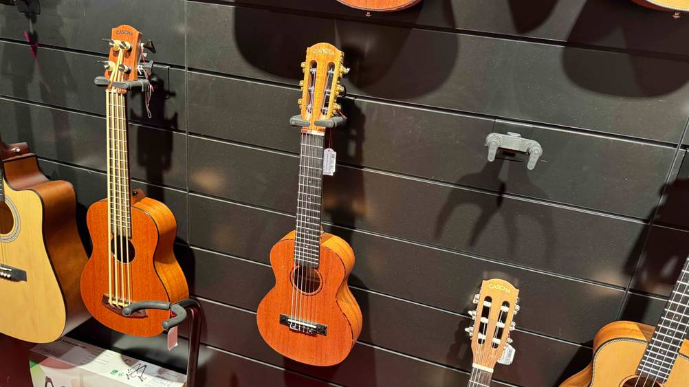 Eine getestete Guitarlele von leicht seitlich mit mehreren anderen Insturmenten an einer schwaren Wand hängend.
