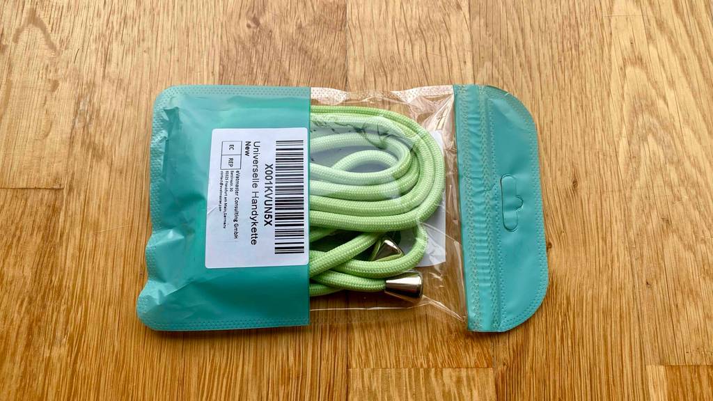Eine verlängerbare Handykette im Test in Grün liegt in einer grün-transparenten Kunststofftüte inklusive Barcode auf einem Holzuntergrund.
