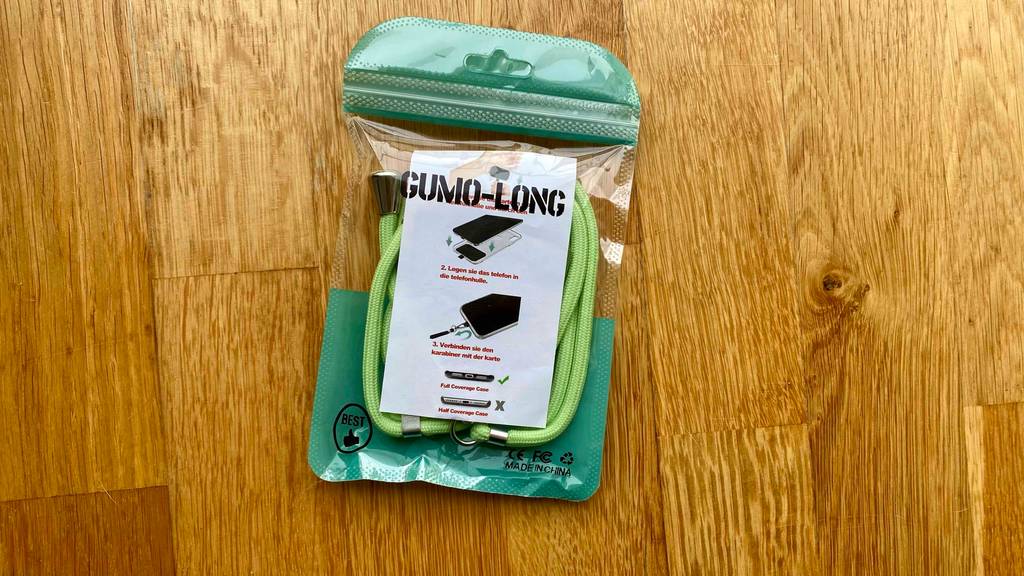 Eine grüne, verlängerbare Handykette im Test befindet sich in seiner transparenten Kunststofftüte inklusive Produktinformationen auf einem Holzuntergrund.