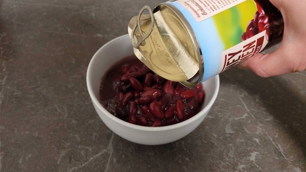Eine Hand schüttet getestete Kidneybohnen aus einer Dose in eine weiße Schale.