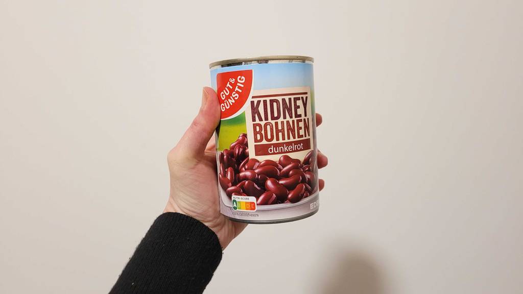 Eine Hand hält getestete Kidneybohnen in einer Dose der Marke Gut & Günstig vor einer weißen Wand.