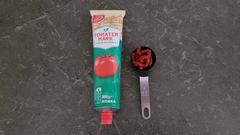 Ein Messlöffel mit Tomatenmark liegt neben einer getesteten Tomatenmark-Tube auf einer dunkelgrauen Fläche.