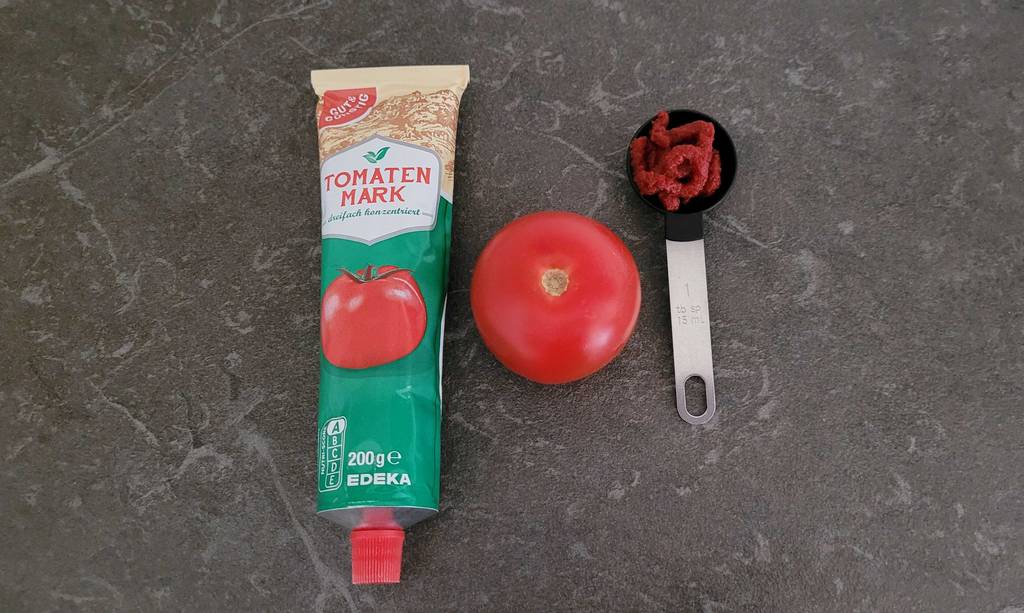 Ein Messlöffel mit getestetem Tomatenmark liegt neben einer frischen Tomate und einer Tube mit Tomatenmark im Test, auf einer grauen Oberfläche.