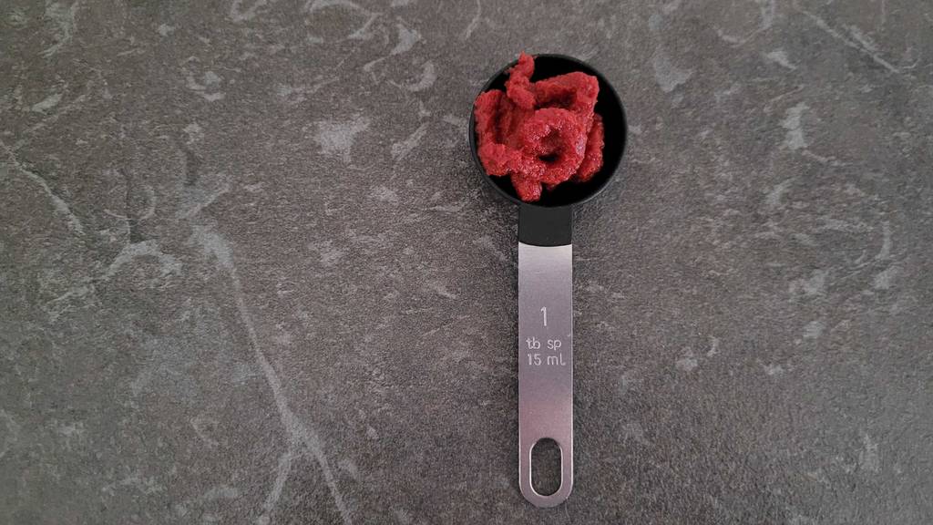 Ein Messlöffel mit getestetem Tomatenmark liegt auf einer dunkelgrauen Oberfläche.