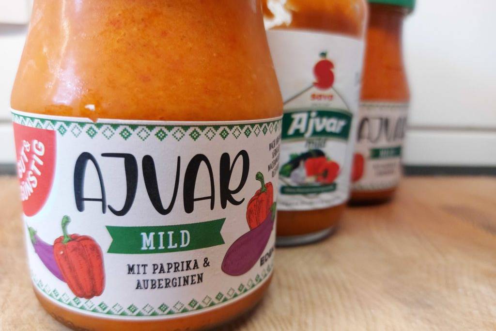 Drei verschiedene Gläser Ajvar zum testen stehen hintereinander auf einer Holz-Oberfläche.