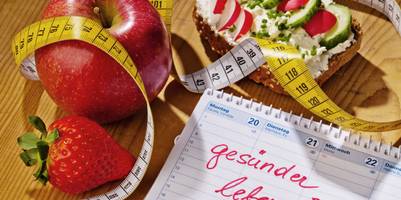 Auf einem Tisch liegt ein Tischkalender, in den in großer Schrift "gesünder leben!" eingetragen wurden. Daneben liegen 1 Apfel, 1 Erdbeere, ein Maßband und ein belegtes Brot mit Gemüsestücken.