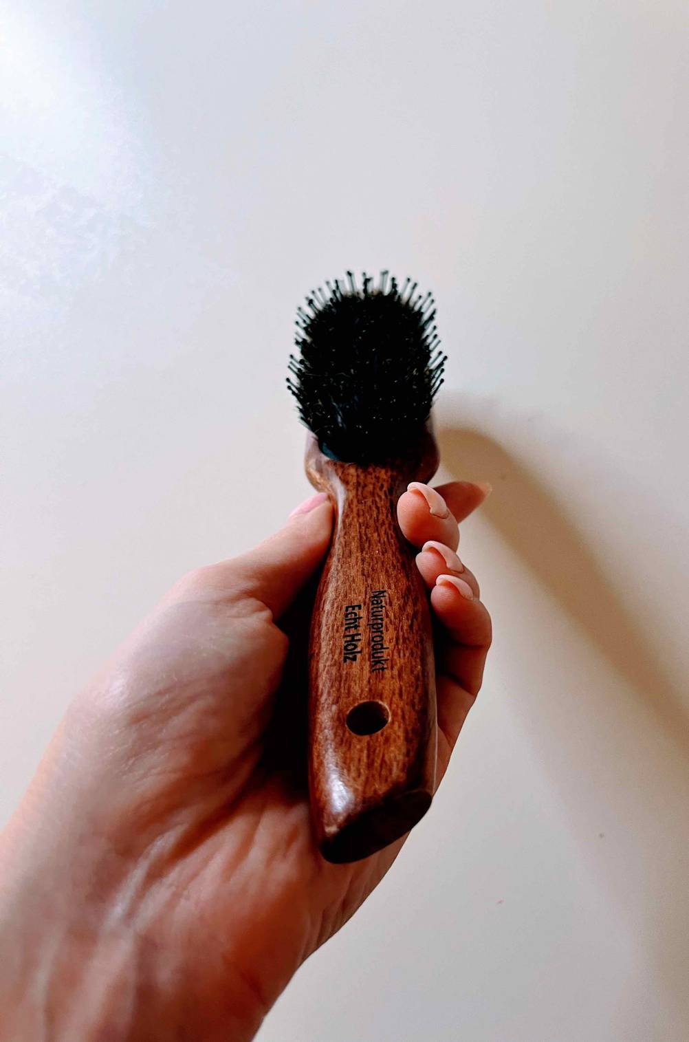 Eine Hand hält eine Haarbürste-Wildschweinborsten vor eine helle Wand.