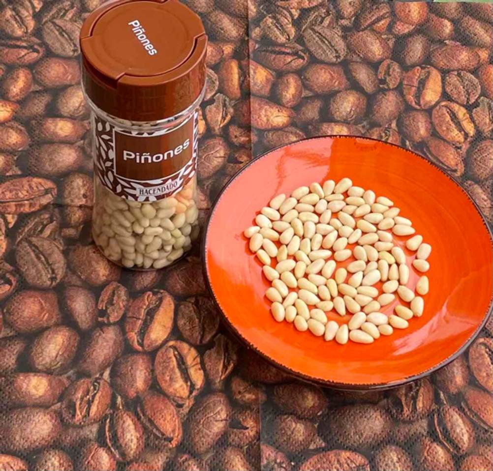 Im Test: eine Flasche Pinienkerne von Hacendado steht neben einer braunen mit Pinienkernen gefüllten Schale auf Tischdecke mit Kaffeebohnenmotiv.