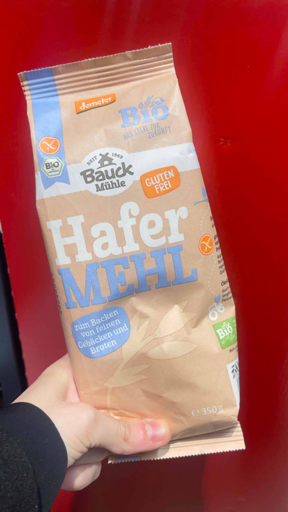 Eine Hand hält die 350-g-Packung mit dem glutenfreien Demeter Hafermehl im Test vor einem roten Hintergrund.