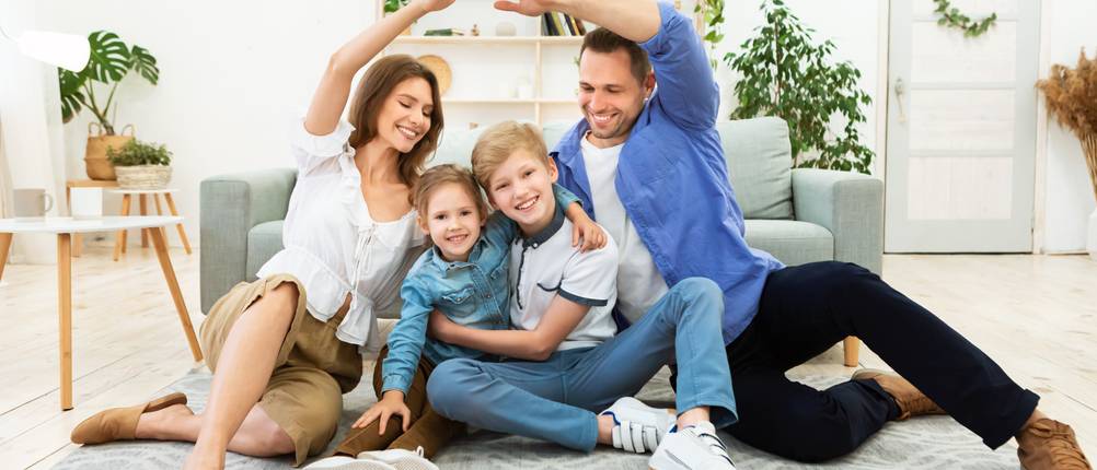 Die Eltern einer Familie formen mit ihren Armen ein Dach über den beiden Kindern, während die gesamte Familie auf dem Boden sitzt.