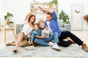 Haftplichtversicherung-Test: Die Eltern einer Familie formen mit ihren Armen ein Dach über den beiden Kindern, während die gesamte Familie auf dem Boden sitzt.
