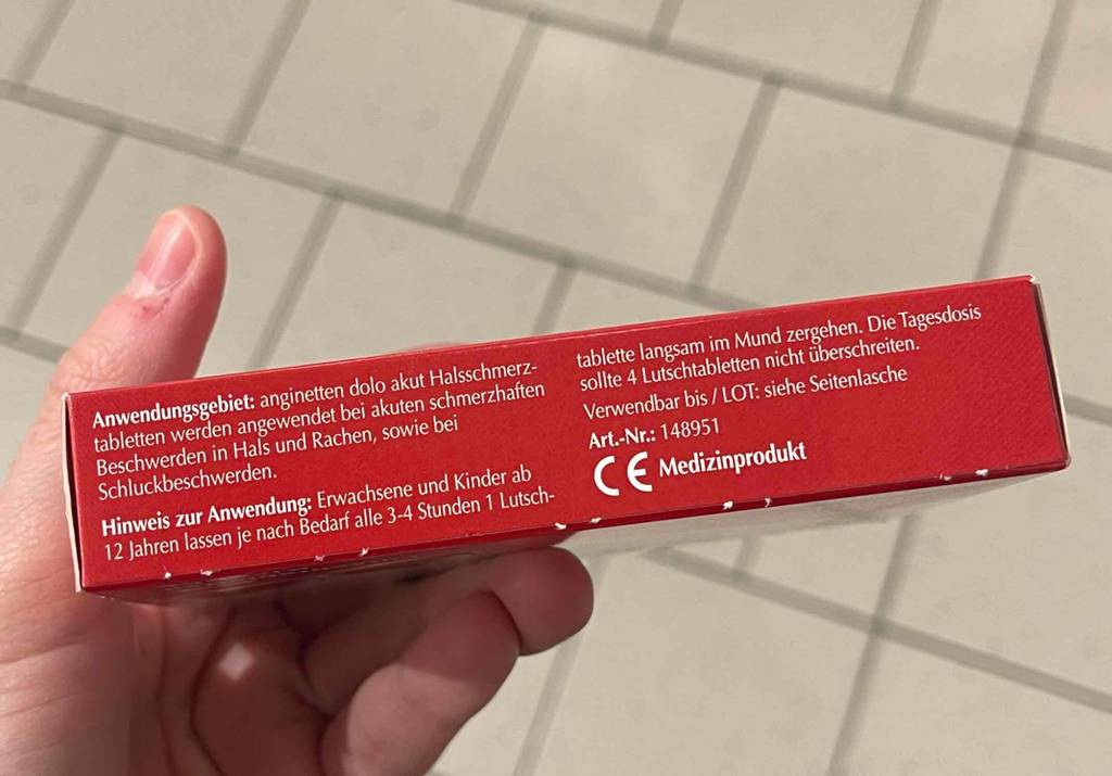 Im Test: Halsschmerztabletten in einer roten Verpackung, inklusive der Anwendungsbeschreibung, in einer Hand über einem weißen Fliesenboden.