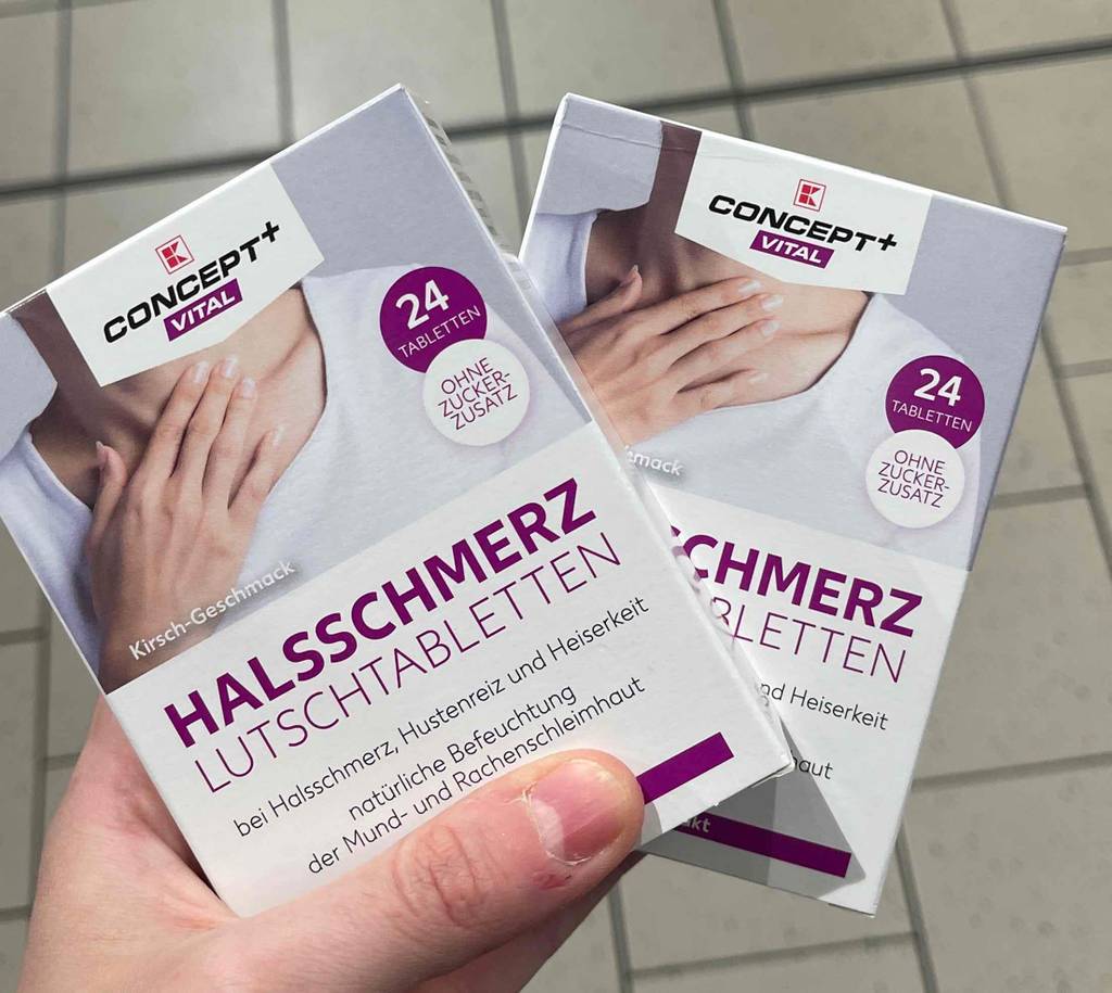 Eine Hand hält zwei 24-Stück-Packungen mit Halsschmerztabletten im Test über einem weißen Fliesenboden.