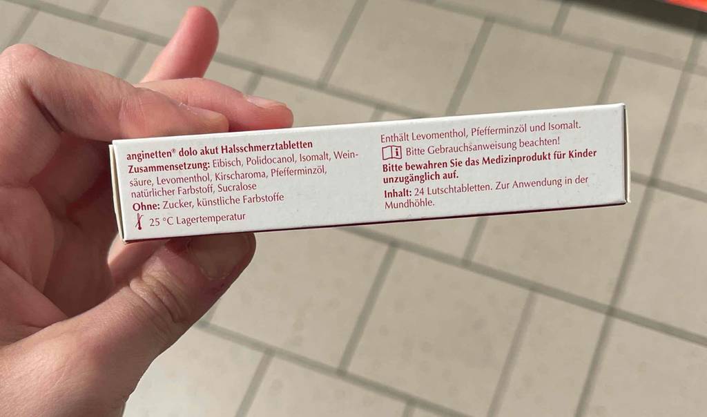Die getesteten Halsschmerztabletten befinden sich innerhalb der Verpackung, mit der gedruckten Zusammensetzung von anginetten-Halsschmerztabletten, die in einer Hand gehalten wird.