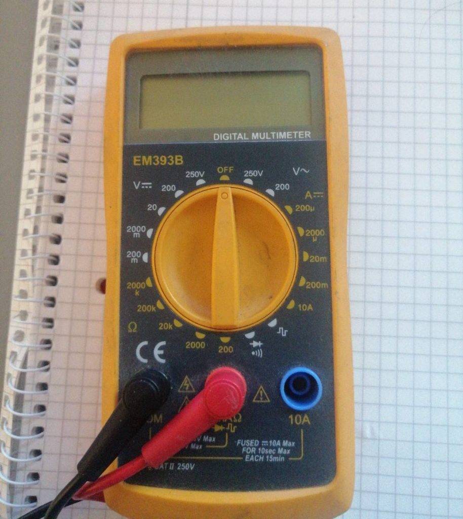 Ein digitaler getesteter Multimeter EM393B von Hama in gelb-blauem Design mit eingestecktem schwarzen und roten Messkabel auf einem Spiral-Rechenheft.